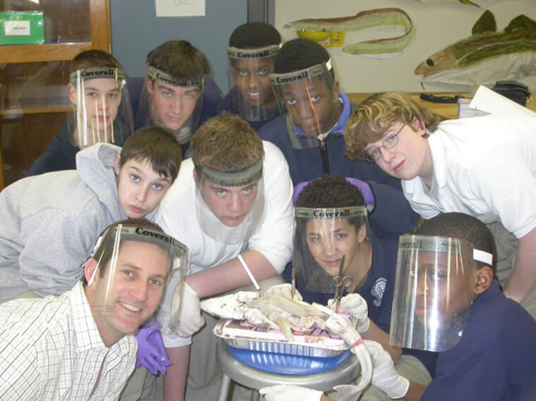 The image shows a group of students and a teacher gathered around a dissection. They are all wearing face shields and gloves. The students appear to be dissecting something on a tray. The setting looks like a science lab.